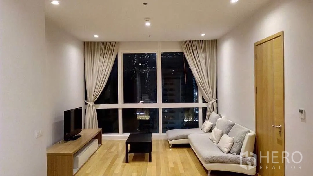 Condominium for rent or sale in Khlong Toei, Bangkok - Bright living room with L-shaped sofa, TV console and floor-to-ceiling windows showing city night views.