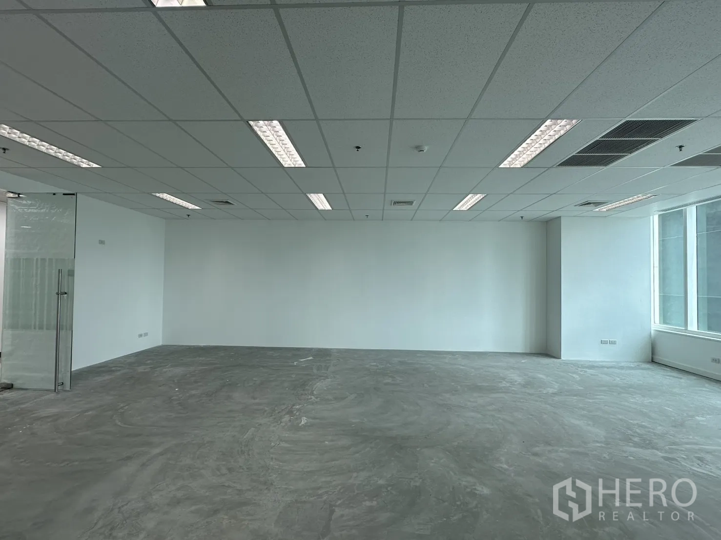 办公室 for rent in Watthana, Bangkok - 宽敞未配家具的办公区，左侧玻璃门、格栅吊顶与混凝土地面。