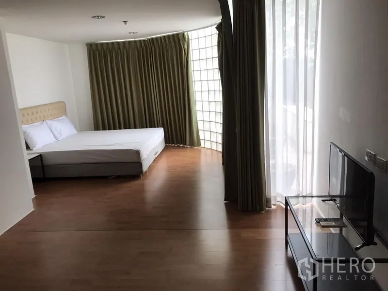 Condominium for rent in Watthana, Bangkok - Large bedroom with queen bed, TV, curved glass‑block wall and sheer curtains allowing natural light.
