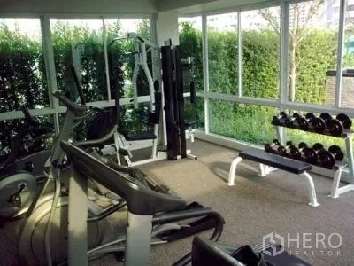 Condominium for rent or sale in Khlong Toei, Bangkok - Condo fitness room with weight machines and dumbbell rack beside garden-view windows.