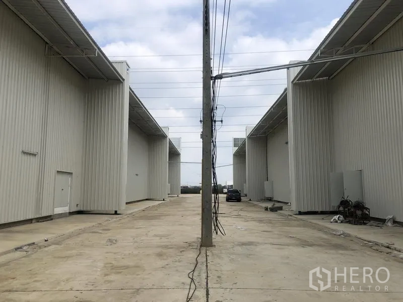 Factory for rent in Khlong Luang, Pathum Thani - Wide access road between factory blocks with a central utility pole and parking areas for trucks.