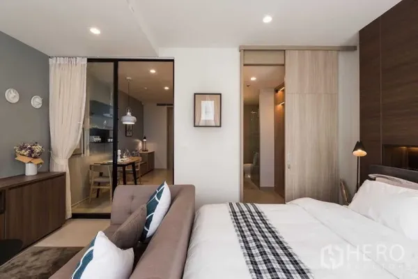 Condominium for rent in Pathum Wan, Bangkok - Modern bedroom with sofa, wood wardrobe and a glass partition opening to the kitchen and dining area.