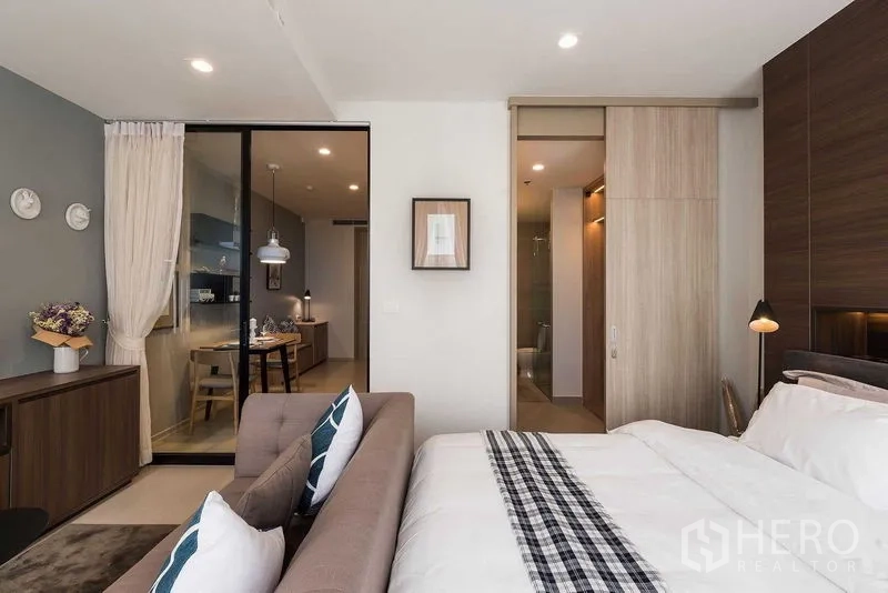 Condominium for rent in Pathum Wan, Bangkok - Modern bedroom with sofa, wood wardrobe and a glass partition opening to the kitchen and dining area.
