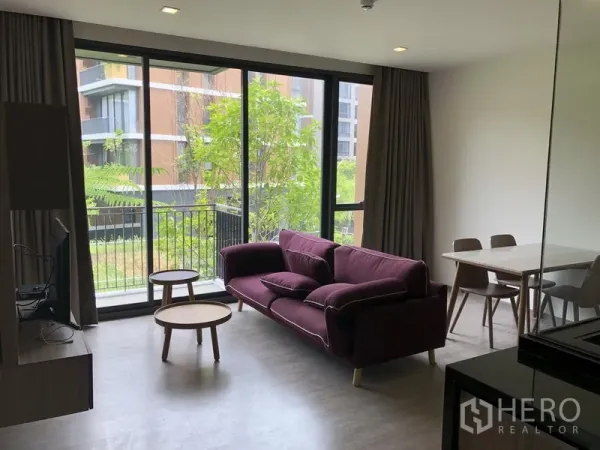 公寓 出租在Watthana, Bangkok - 采光充足的客厅，落地窗与阳台望向绿意，紫色沙发与茶几、餐桌一角