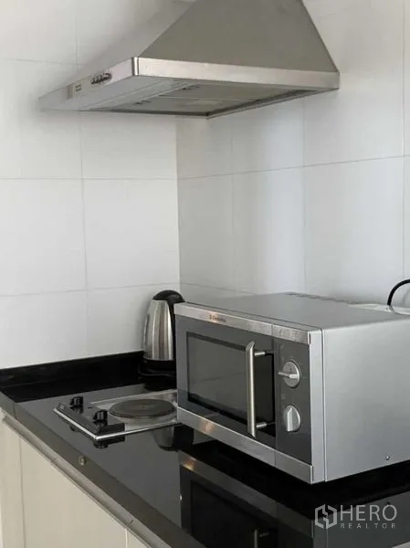 Condominium for rent in Khlong Toei, Bangkok - Compact kitchen with black granite countertop, electric cooktop, microwave, kettle, and stainless steel range hood.