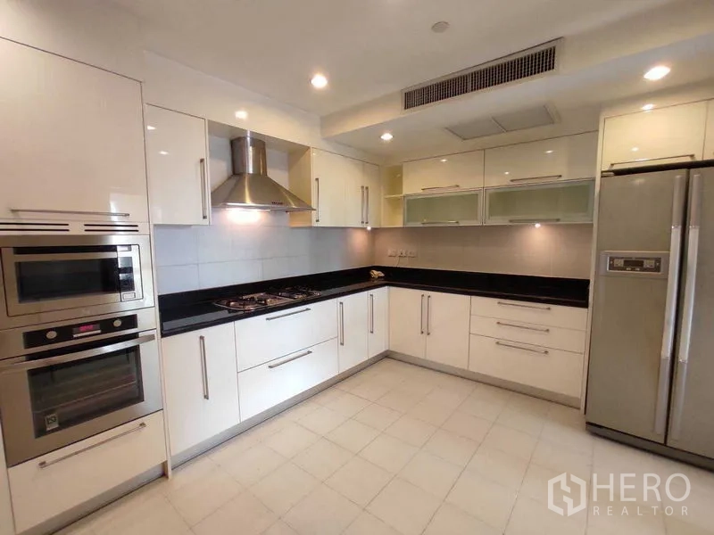 Condominium for rent in Khlong Toei, Bangkok - Modern U‑shaped kitchen with white cabinetry, gas hob, hood, wall ovens and a double‑door refrigerator.