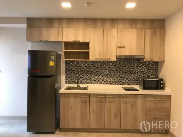 Condominium for rent in Watthana, Bangkok - Fitted kitchen with wood cabinetry, mosaic tile backsplash, cooktop, sink, microwave, and refrigerator.