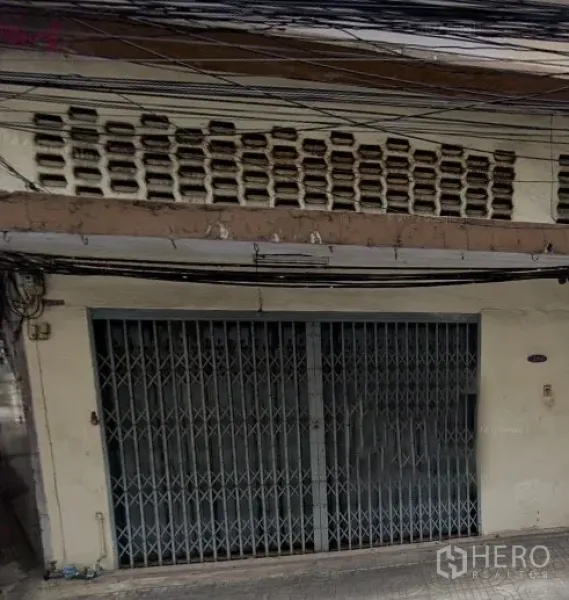 Shophouse for rent in Khlong Toei, Bangkok - Street-front shophouse exterior in Khlong Toei with wide metal folding gate, ventilation blocks, overhead utility cables and a covered sidewalk.