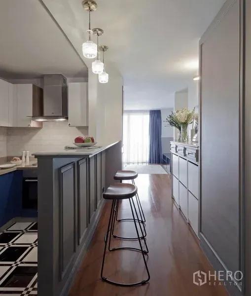Condominium for sale in Watthana, Bangkok - Galley kitchen with breakfast bar, pendant lights and a corridor leading to a bright balcony.