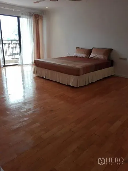 Condominium for rent in Khlong Toei, Bangkok - Spacious master bedroom with wood floors, bed, and sliding doors to a balcony with light curtains.