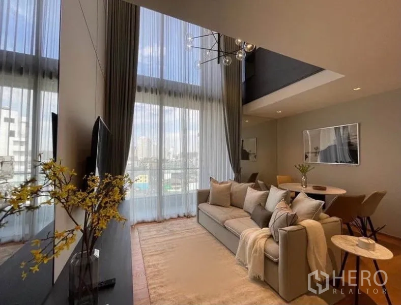 Condominium for rent in Khlong Toei, Bangkok - Sunlit lounge with neutral sofa, large rug and round dining table under double-height glass.