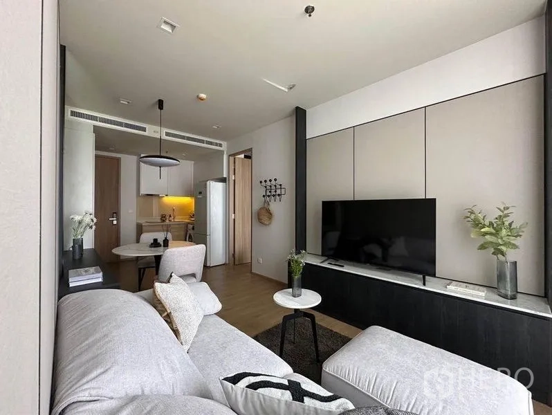 公寓 出租在Watthana, Bangkok - 电视墙搭配浅灰组合沙发与小圆几，可见厨房入口。