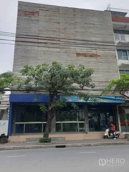 Retail Space for rent in Watthana, Bangkok - Street-facing retail building with a full-width glass storefront, blue awning, and mature tree along the sidewalk.
