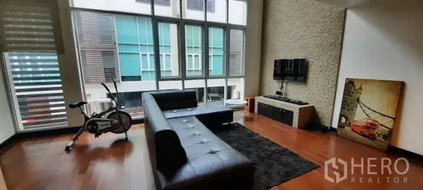Townhouse for rent or sale in Khlong Toei, Bangkok - High-ceiling living room with floor-to-ceiling windows, black sectional sofa, TV feature wall, and exercise bike.