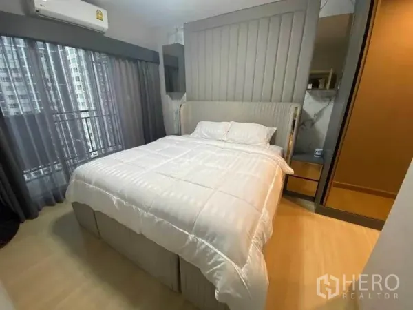 Condominium for rent or sale in Huai Khwang, Bangkok - Modern condo bedroom in Huai Khwang, Bangkok with padded headboard, white bedding, mirrored wardrobe, air conditioner, and floor-to-ceiling window with city view.