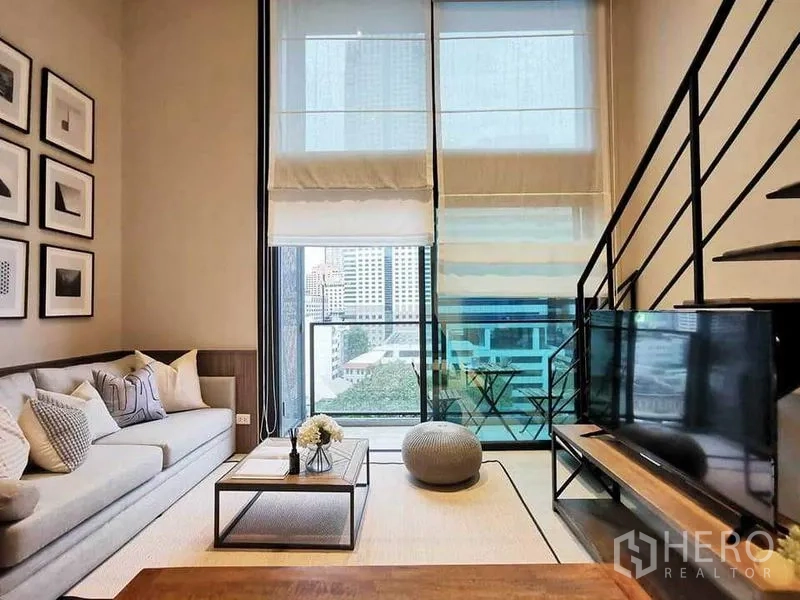 Condominium for rent in Bang Rak, Bangkok - Living room facing a balcony with tall windows, city skyline and black metal stairs.