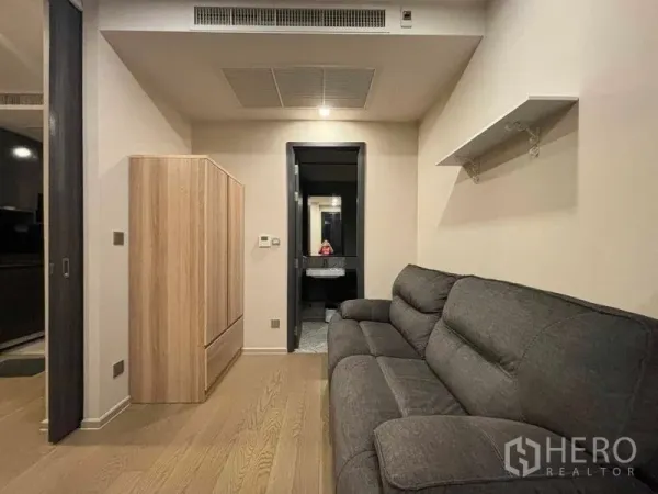 Condominium for rent in Watthana, Bangkok - Cozy lounge with gray sofa, wardrobe and a view toward the modern bathroom with vanity.