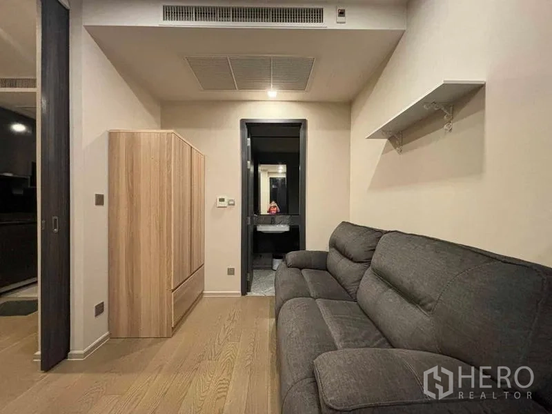 Condominium for rent in Watthana, Bangkok - Cozy lounge with gray sofa, wardrobe and a view toward the modern bathroom with vanity.