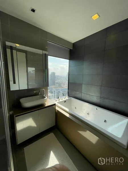 Condominium for rent or sale in Bang Rak, Bangkok - Bathroom with a large jetted bathtub by the window overlooking the Bangkok skyline and a sleek vanity.