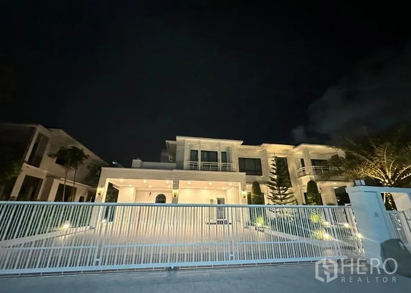 独立式住宅 出租在Bang Phli, Samut Prakan - 夜景下透过白色金属栅栏看到灯光辉映的现代住宅。