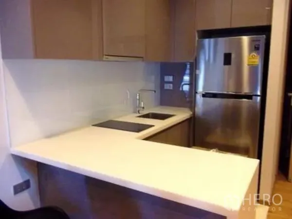 Condominium for rent or sale in Watthana, Bangkok - L-shaped kitchenette with white countertop, sink, cooktop and stainless-steel refrigerator.