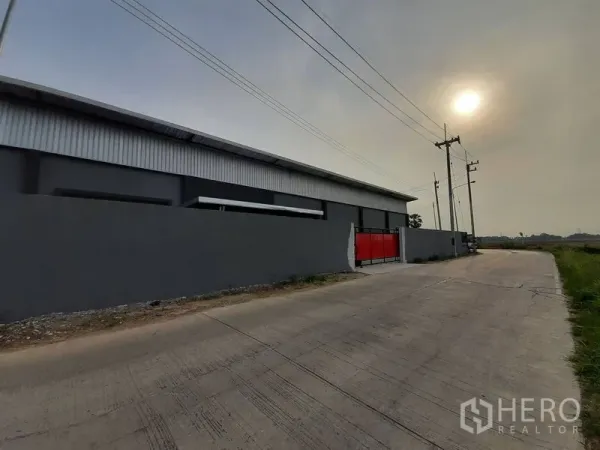Warehouse for rent in Lam Luk Ka, Pathum Thani - Street view of the gated warehouse wall and red entrance gate along a concrete road.