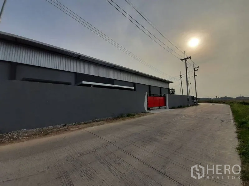 Warehouse for rent in Lam Luk Ka, Pathum Thani - Street view of the gated warehouse wall and red entrance gate along a concrete road.
