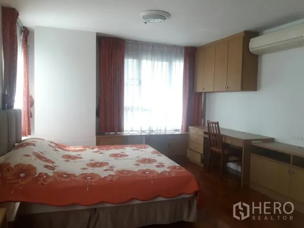 Condominium for rent in Khlong Toei, Bangkok - Bright primary bedroom with large windows, built-in desk and storage, air conditioning and a floral bedspread.