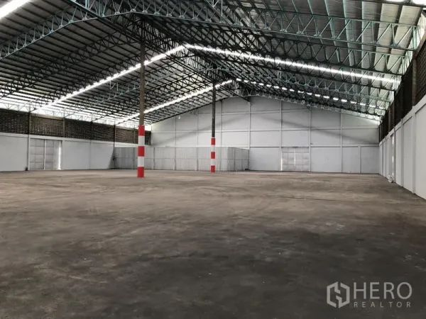 Warehouse for rent in Mueang Pathum Thani, Pathum Thani - Spacious warehouse interior with high steel truss roof, concrete floor, and red‑white marked columns.