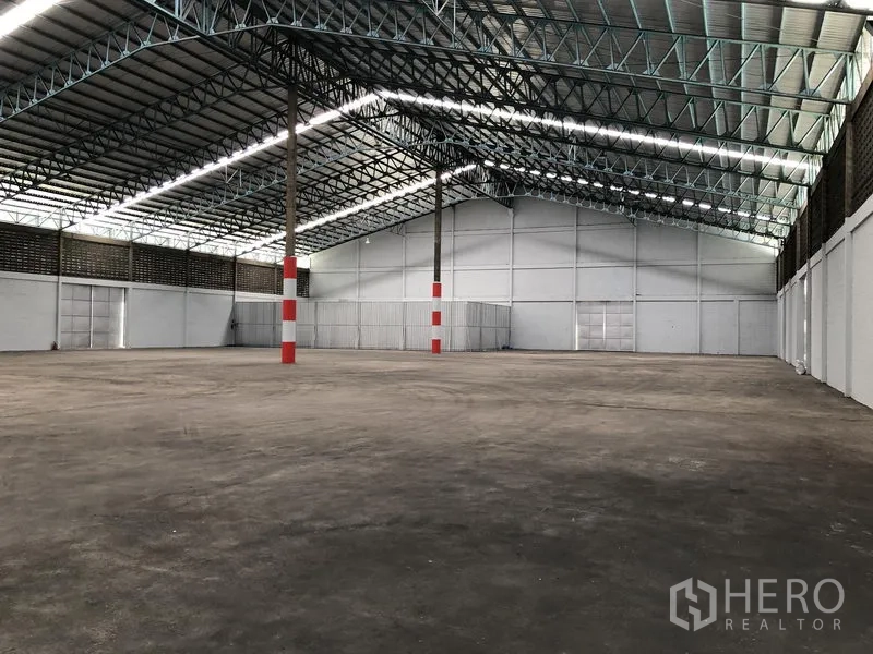 Warehouse for rent in Mueang Pathum Thani, Pathum Thani - Spacious warehouse interior with high steel truss roof, concrete floor, and red‑white marked columns.