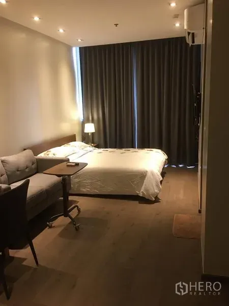 公寓 出租在Khlong Toei, Bangkok - 开放式卧室与客厅，双人床、灰色遮光窗帘、沙发与边桌，嵌灯照明。