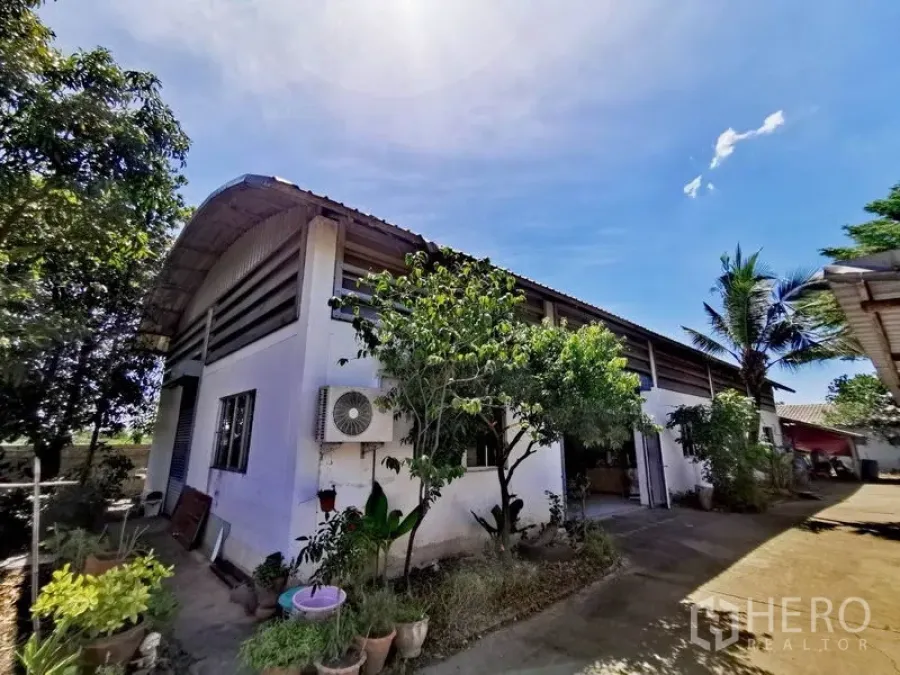 货栈 出售在Pathum Thani - 仓库外观，弧形金属屋顶与白色墙面，绿植与车道，蓝天之下。