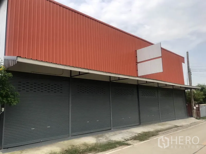 Warehouse for rent in Lam Luk Ka, Pathum Thani - Street-front orange metal-clad warehouse with multiple gray rolling shutters and a covered loading awning.