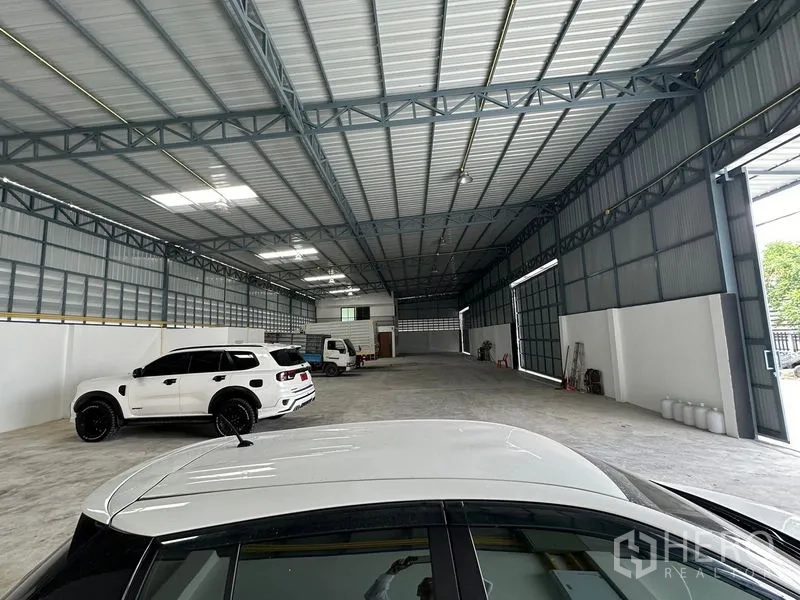 Warehouse for rent in Lam Luk Ka, Pathum Thani - Deep view across the warehouse with steel roof, side louvers and vehicles on the floor.
