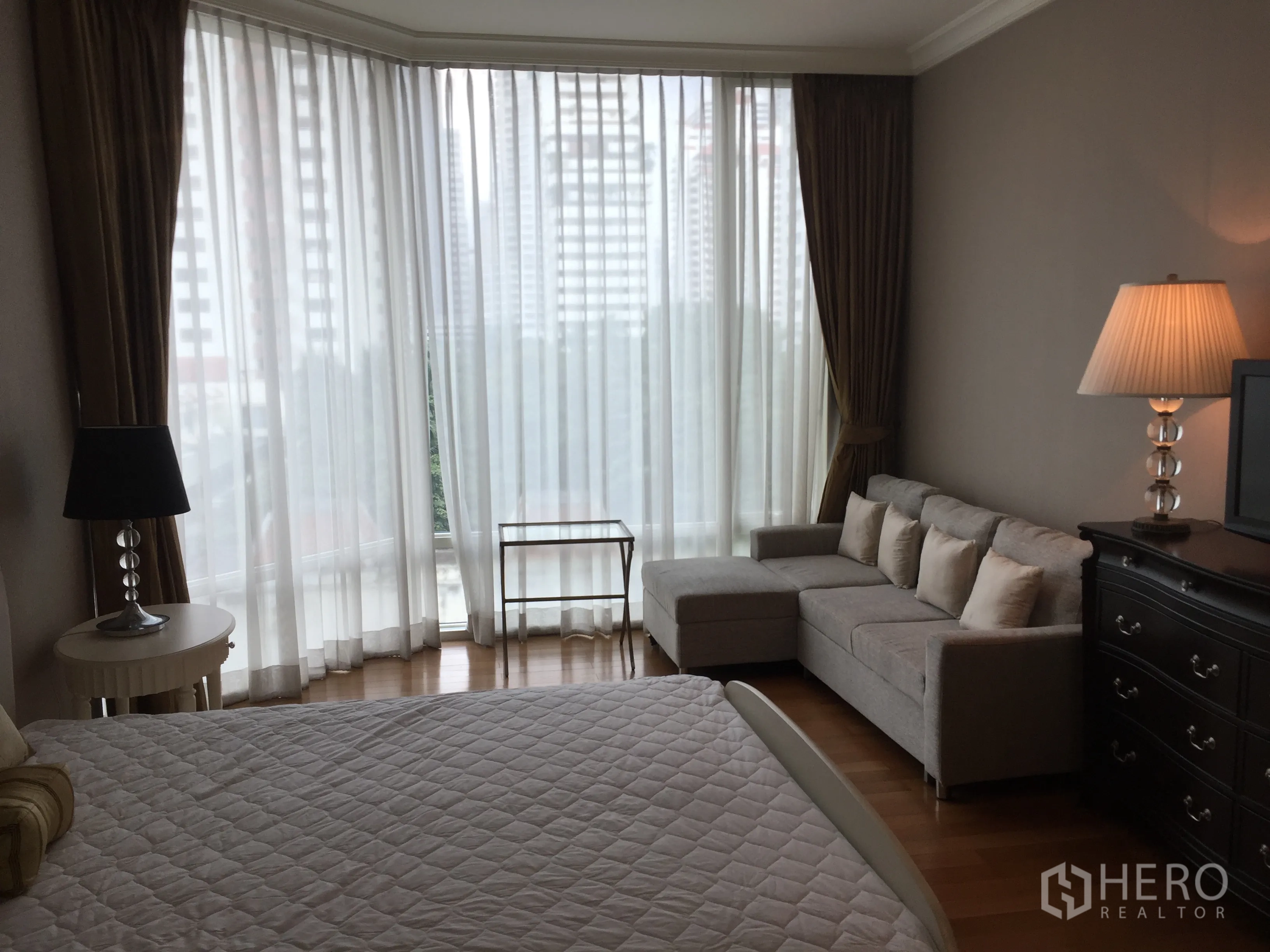 Condominium for rent in Watthana, Bangkok - Corner bedroom with sheer curtains, L-shaped sofa and wide city view windows.