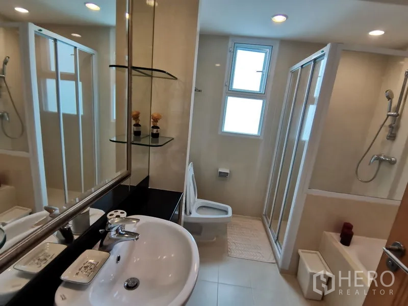 Condominium for rent in Khlong Toei, Bangkok - Light tiled bathroom with vanity, glass-enclosed shower, and separate bathtub.