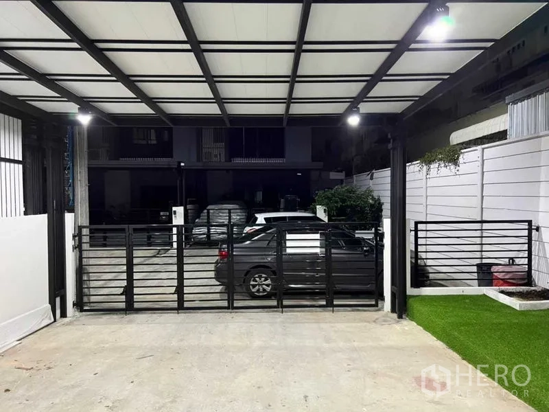 联排别墅 出租在Wang Thonglang, Bangkok - 带顶棚双车位车库，金属大门、照明与一侧人造草坪。