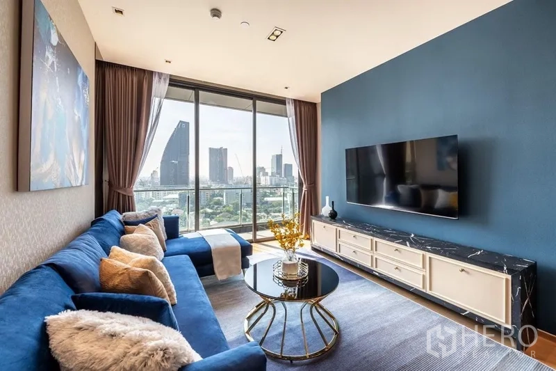 Condominium for rent or sale in Khlong Toei, Bangkok - Bright living room with blue sectional sofa, TV wall, and floor‑to‑ceiling windows opening to a balcony with Bangkok skyline views.