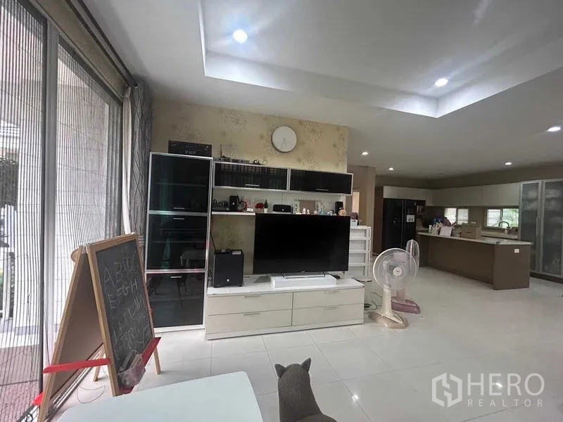 บ้าน ขายในSamut Prakan - ห้องนั่งเล่นสว่างพร้อมชั้นวางทีวี ฝ้าไฟดาวน์ไลท์ และครัวกว้างด้านใน
