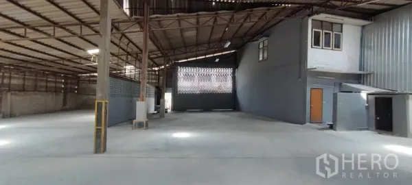 Warehouse for rent in Phra Khanong, Bangkok - Open-plan warehouse interior with high steel truss roof, concrete floor, and an internal mezzanine office beside a large loading gate.