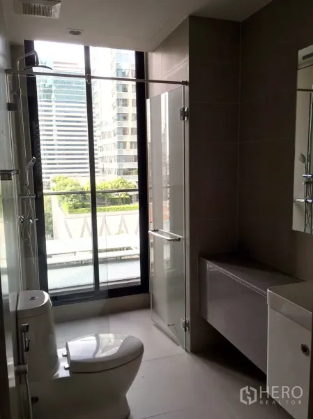 Condominium for rent in Pathum Wan, Bangkok - Modern bathroom with glass shower, toilet, and large window overlooking city buildings.