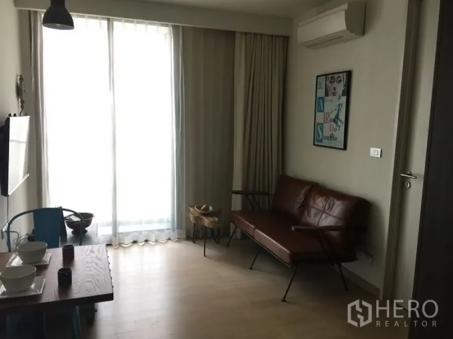 Condominium for rent in Watthana, Bangkok - Bright living room with brown leather sofa, floor-to-ceiling curtains, wall air-conditioner and dining nook.
