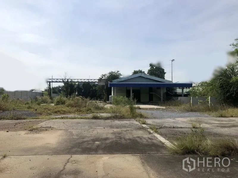 Warehouse for rent or sale in Lam Luk Ka, Pathum Thani - Expansive concrete yard with a small blue-roof office and overgrown edges.