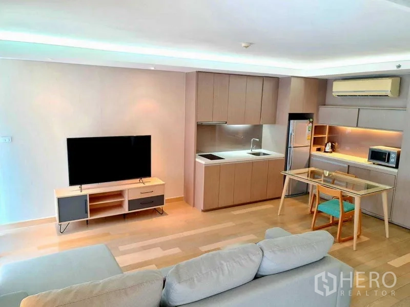 Condominium for rent in Watthana, Bangkok - Open-plan living area with kitchenette, dining table and large TV on light wood flooring.