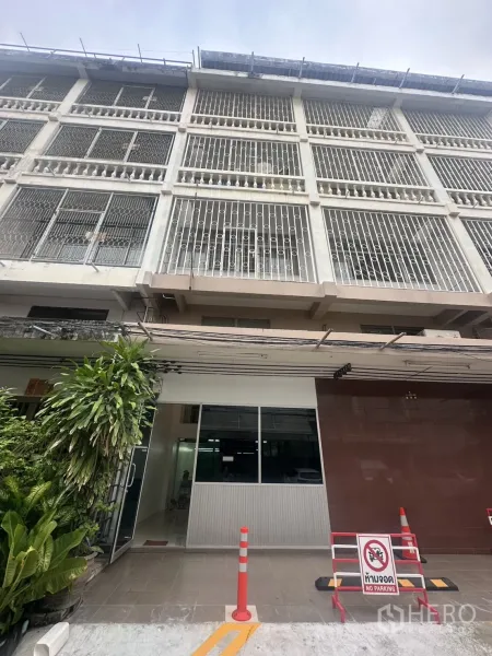 办公空间 出租在Khlong Toei, Bangkok - 多层办公楼外立面，金属护栏与玻璃入口，门前有植物和禁停标志。