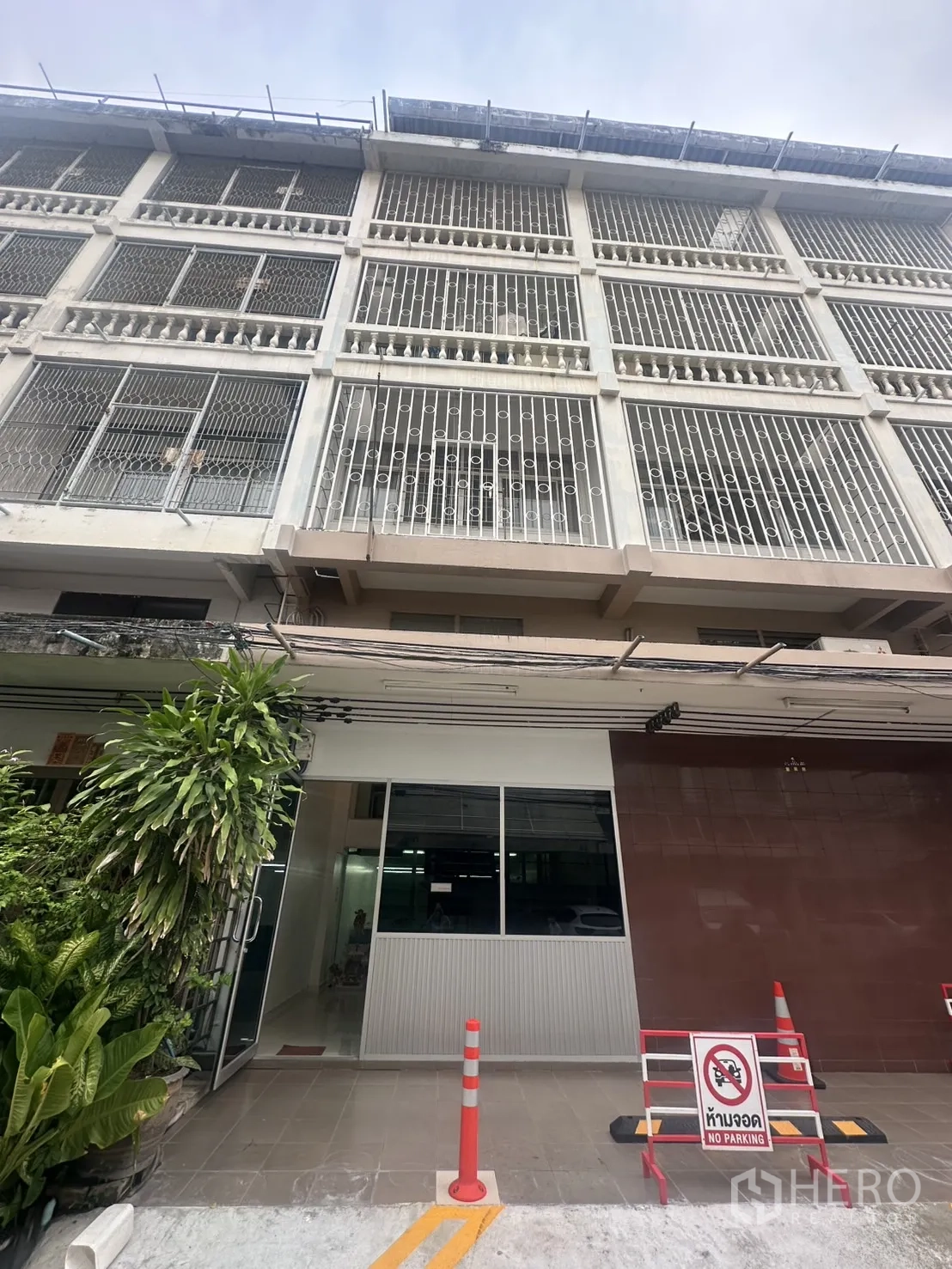 办公空间 出租在Khlong Toei, Bangkok - 多层办公楼外立面，金属护栏与玻璃入口，门前有植物和禁停标志。