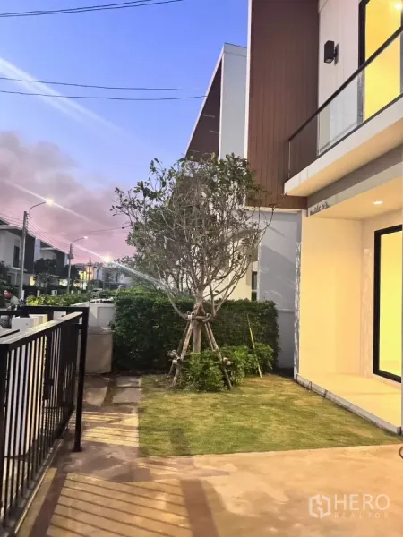 Detached House for rent in Samut Prakan - Front garden with sprinkler, walkway and neighboring modern homes at sunset.