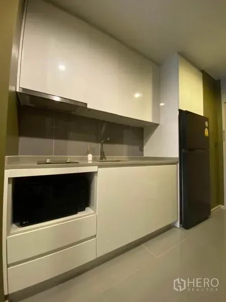 Condominium for rent in Watthana, Bangkok - Compact modern kitchenette with glossy white cabinets, sink, hob and a full-size refrigerator.