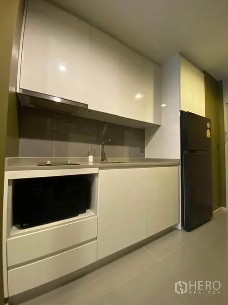 Condominium for rent in Watthana, Bangkok - Compact modern kitchenette with glossy white cabinets, sink, hob and a full-size refrigerator.