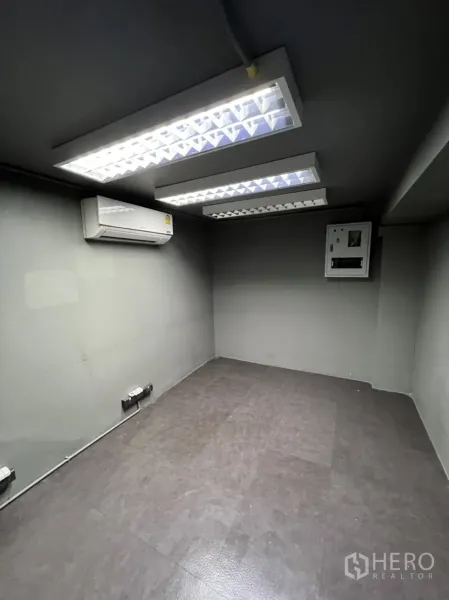 Shophouse for rent in Phra Khanong, Bangkok - Compact office room with fluorescent ceiling lights, split air conditioner and an electrical panel on grey walls.